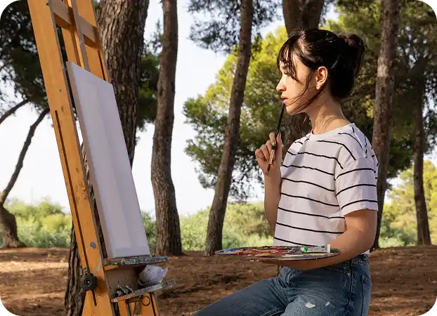 A girl in the park is painting on a canvas on an easel, holding a palette.