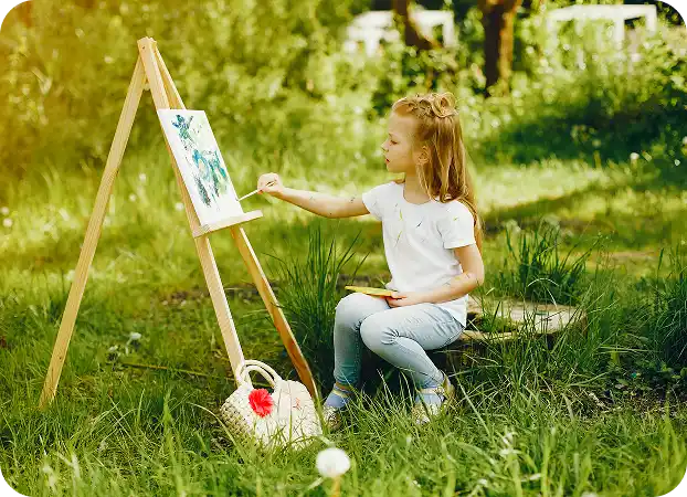 A small girl in the park is painting on a canvas on an easel, holding a palette.
